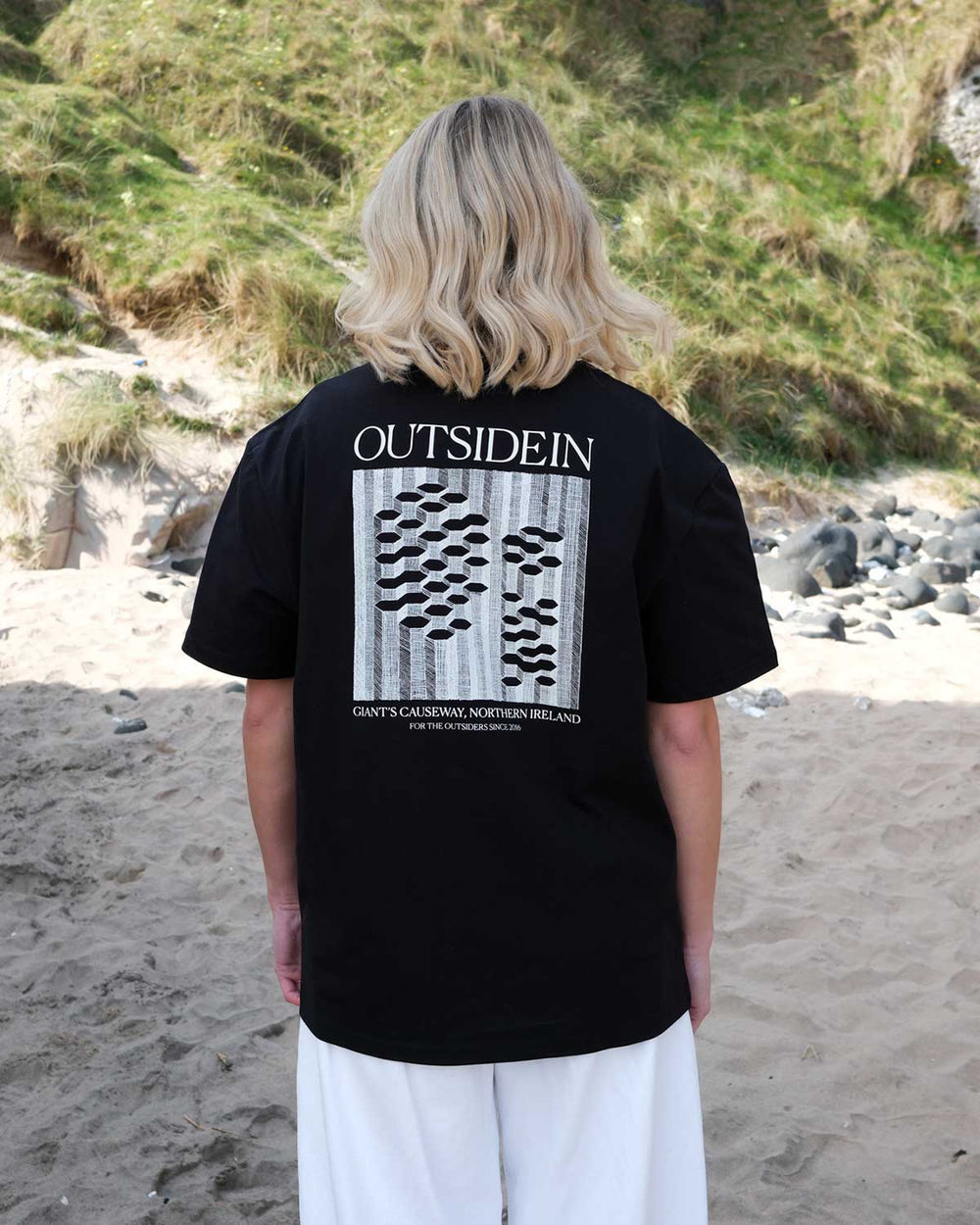 Back view of an OutsideIn Black Abstract Causeway T shirt on a beach