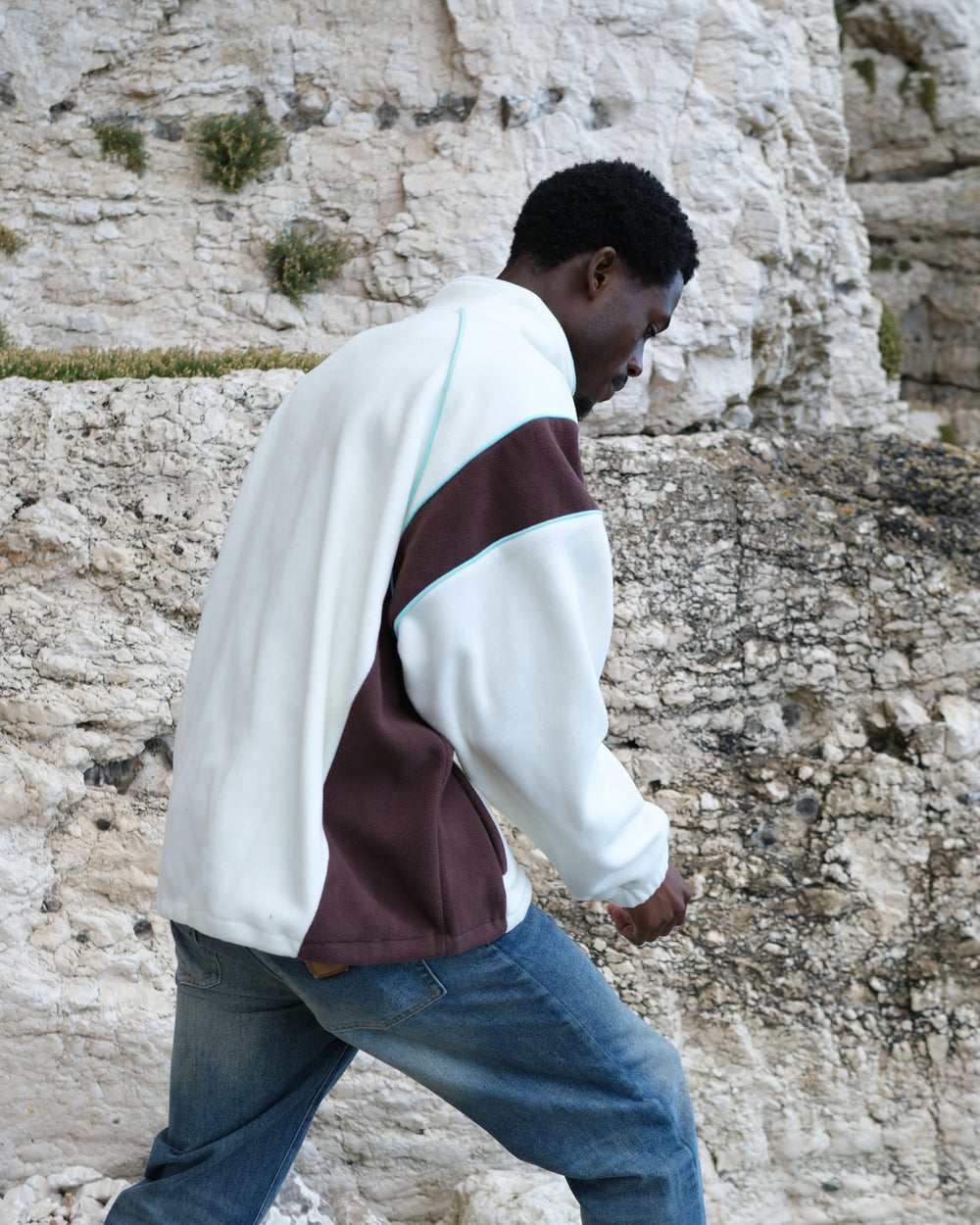 A man wearing an OutsideIn cream fleece jacket with white, dark brown, and light blue accents, paired with blue jeans, walking outdoors against a rocky cliff background.
