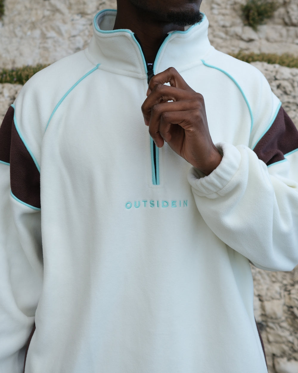 A close-up of a person wearing a white quarter-zip fleece with brown shoulder panels and light blue piping details. The brand name 'OUTSIDEIN' is printed in light blue on the chest.