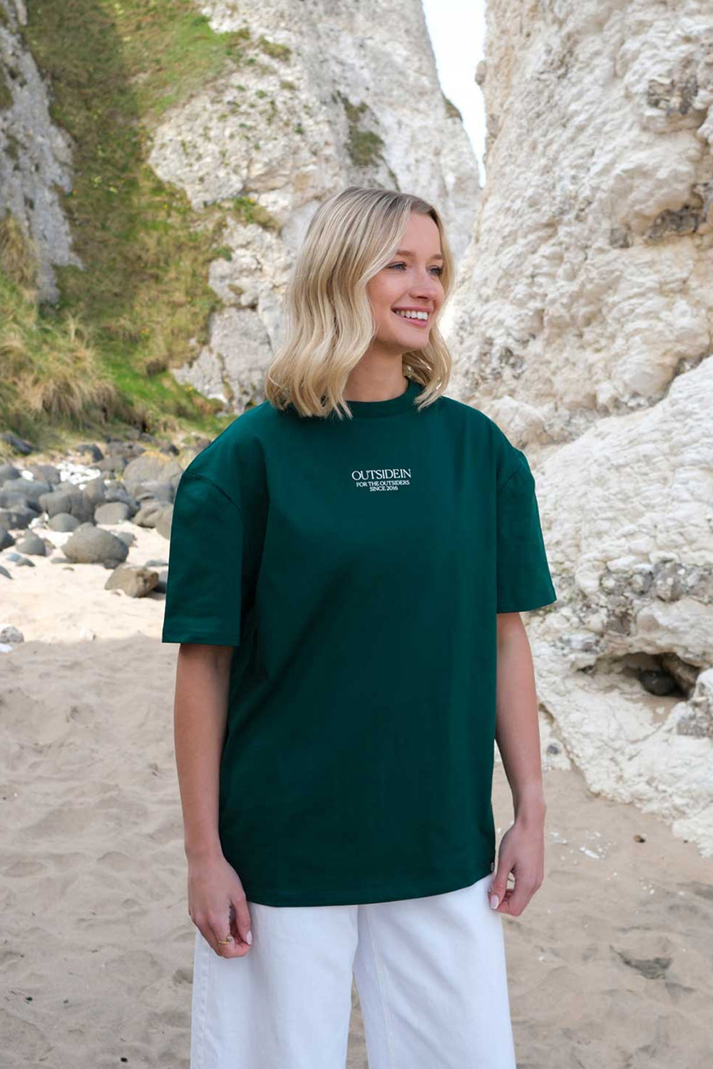 Model wearing a green OutsideIn t-shirt with text on it, standing on a beach.
