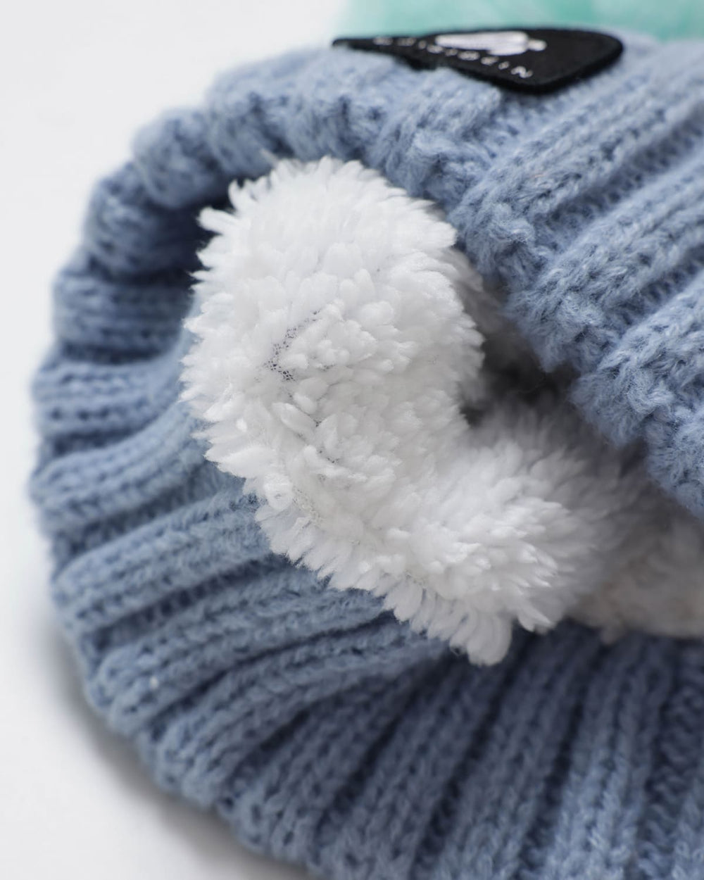 Close-up of OutsideIn light blue ribbed knit pom pom hat with a white faux-fur trim and a black brand patch.