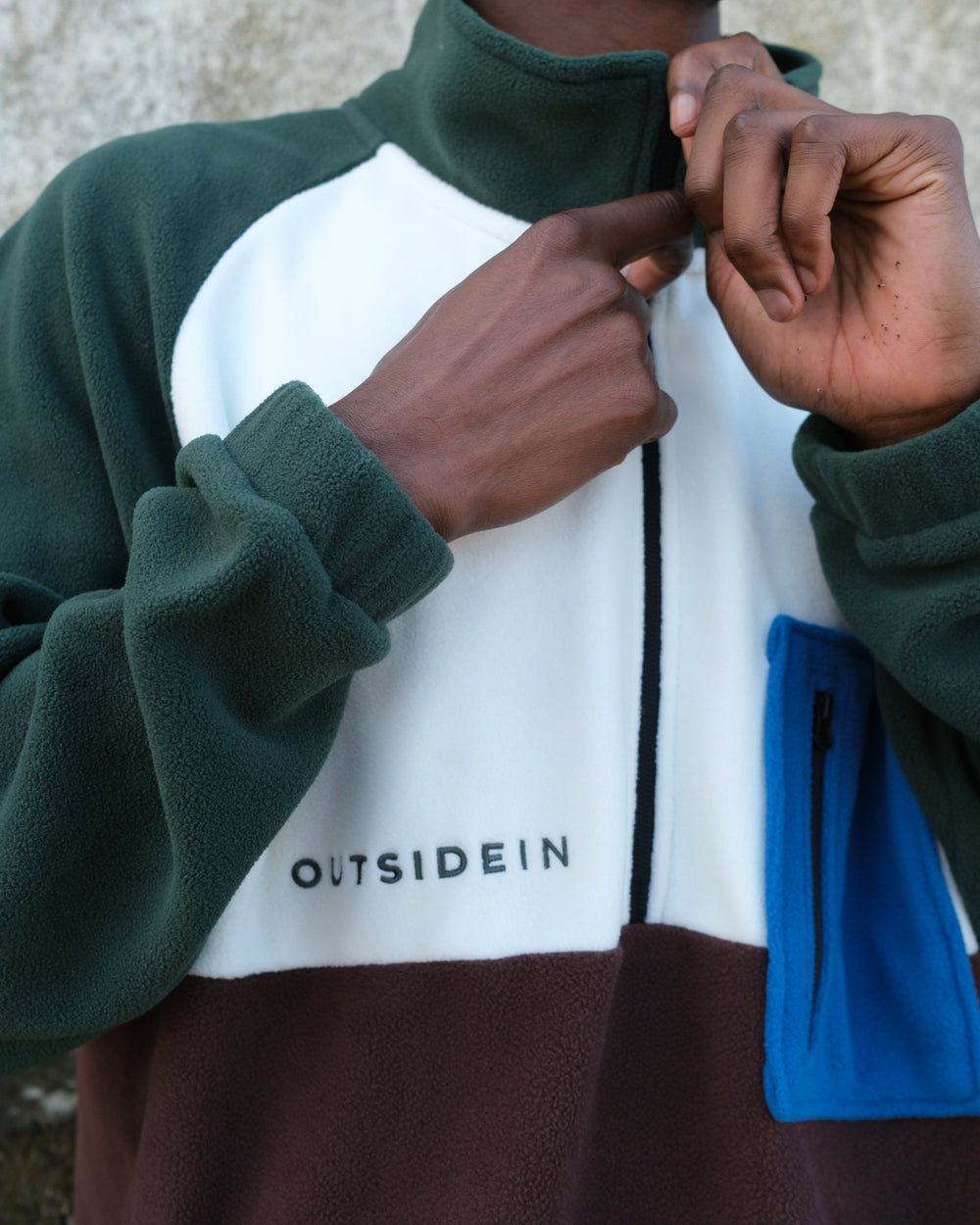 A close-up of a person wearing a colour-block fleece pullover with dark green, white, brown, and blue panels. The person is pulling the zipper with both hands, and the brand 'OUTSIDEIN' is embroidered on the white chest panel.