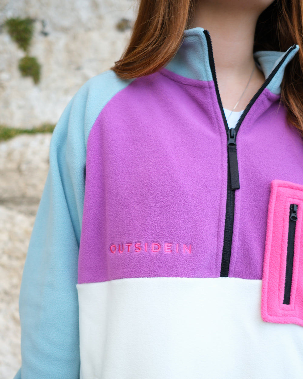 A close-up of a person wearing a colour-blocked quarter-zip fleece pullover. The garment features a light blue left sleeve, a purple right chest panel with 'OUTSIDEIN' embroidered in pink, and a white lower panel with a bright pink zippered chest pocket.
