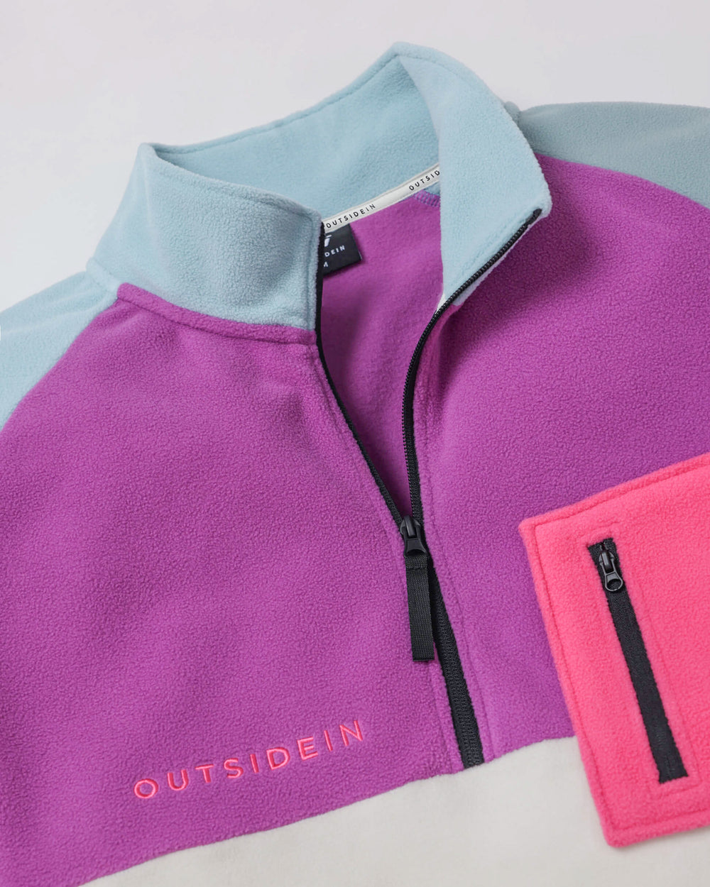 Close-up of a purple and blue fleece with OUTSIDEIN embroidered in pink across the center, with a white fleece section visible below.This is a half zip fleece.