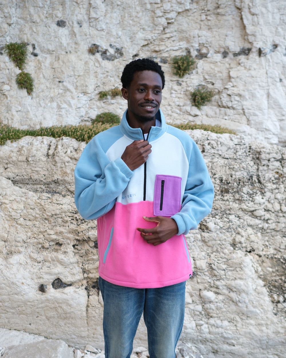 A man wearing a colour-blocked fleece blue and pink sections, standing in front of a textured beige rock cliff.