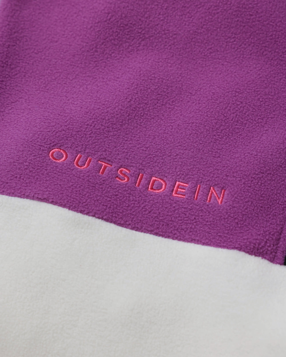 Close-up of a purple fleece fabric with the word 'OUTSIDEIN' embroidered in pink across the center, with a white fleece section visible below.