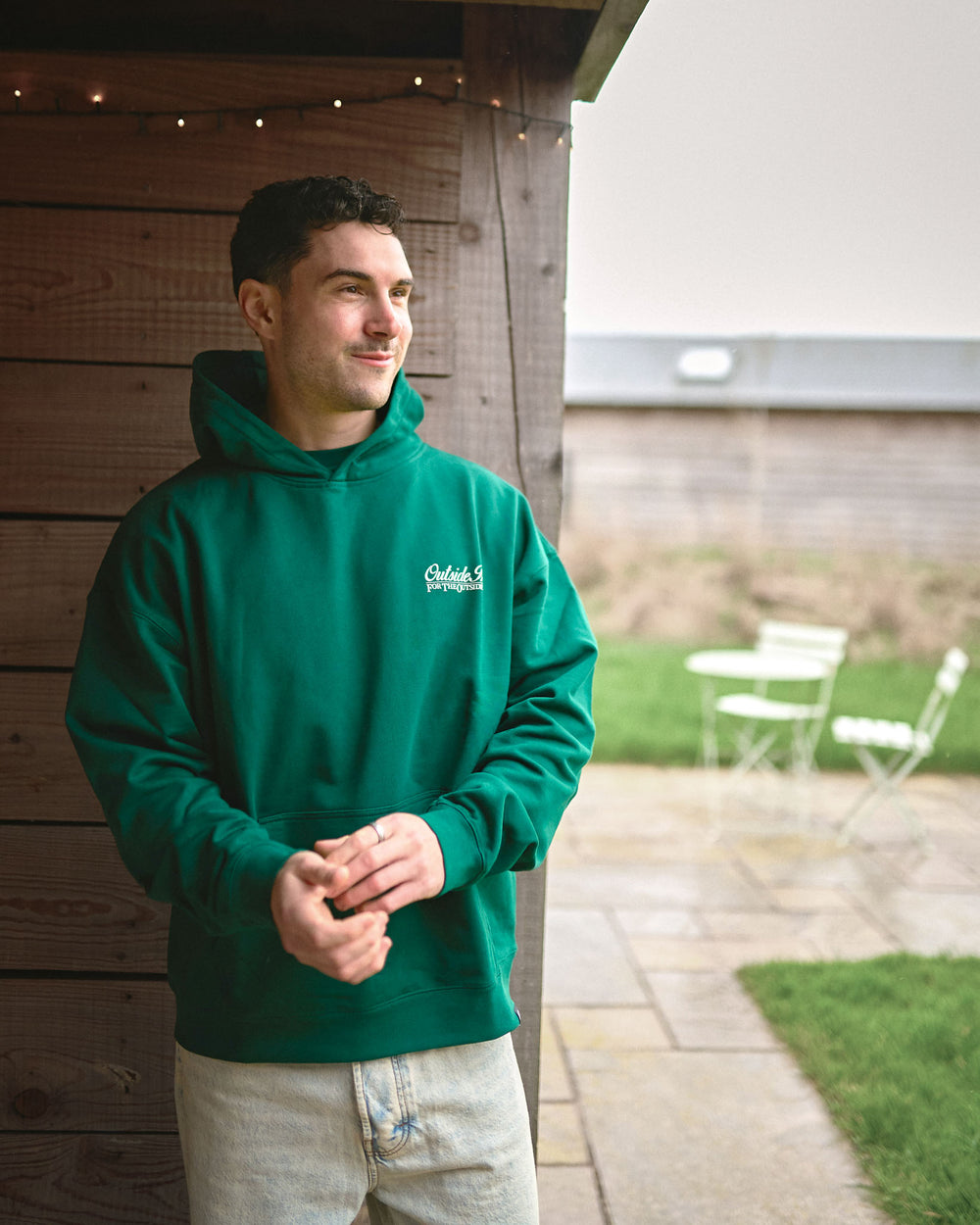 Man wearing a green hoodie with a logo, standing outdoors near a wooden structure.