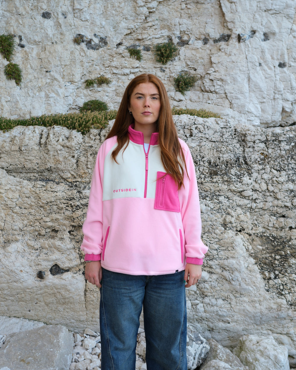 A woman stands outdoors in front of a light-colored rocky cliff wearing a pink and white half-zip fleece pullover with blue jeans.