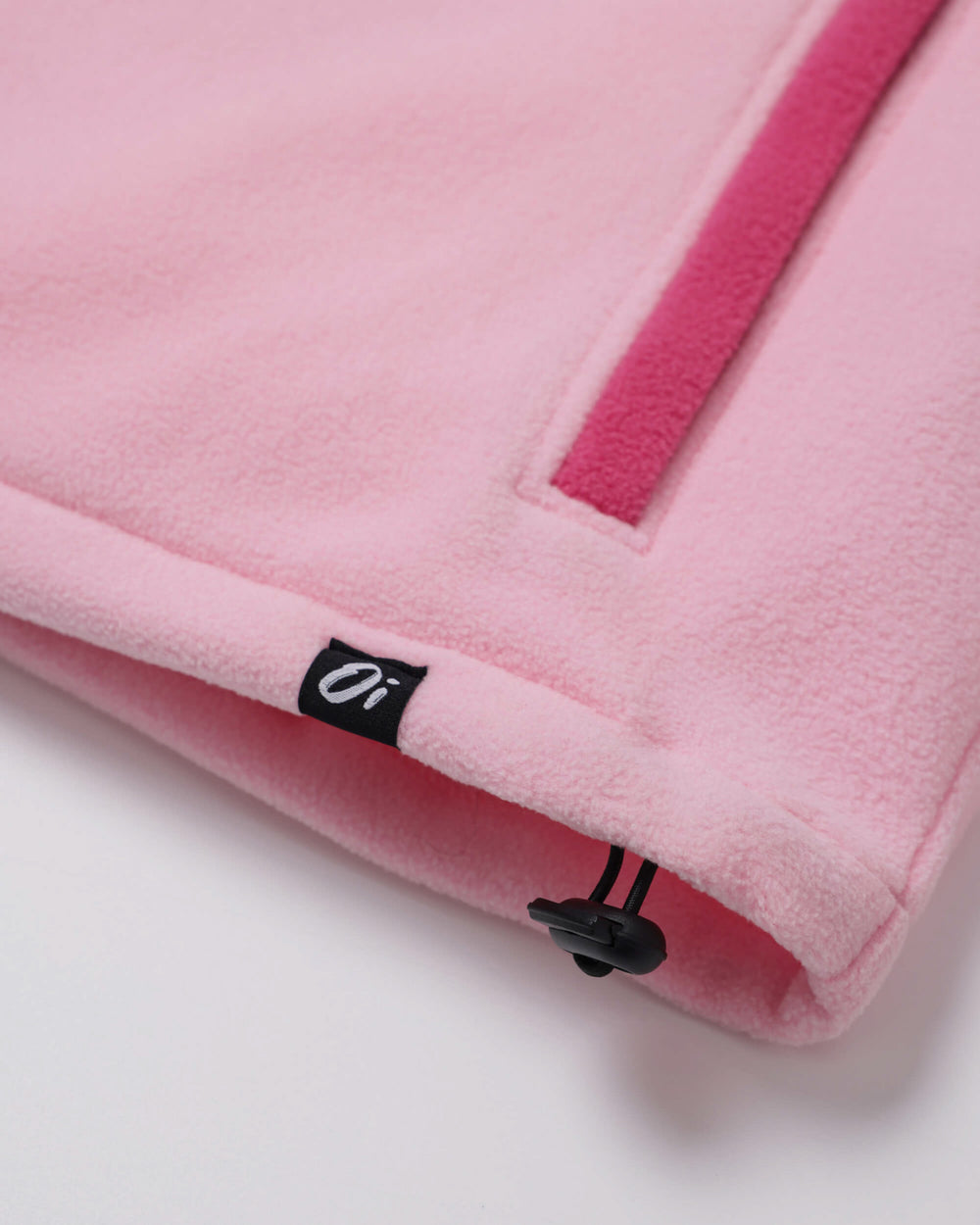 Close-up of a pink fleece garment corner with a black drawstring and a small black brand tag featuring a white stylized logo.