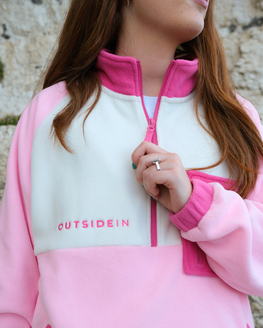 A close-up of a person wearing a pink and white fleece zip-up sweatshirt with the brand name 'OUTSIDEIN' printed on the chest, set against a stone wall background.