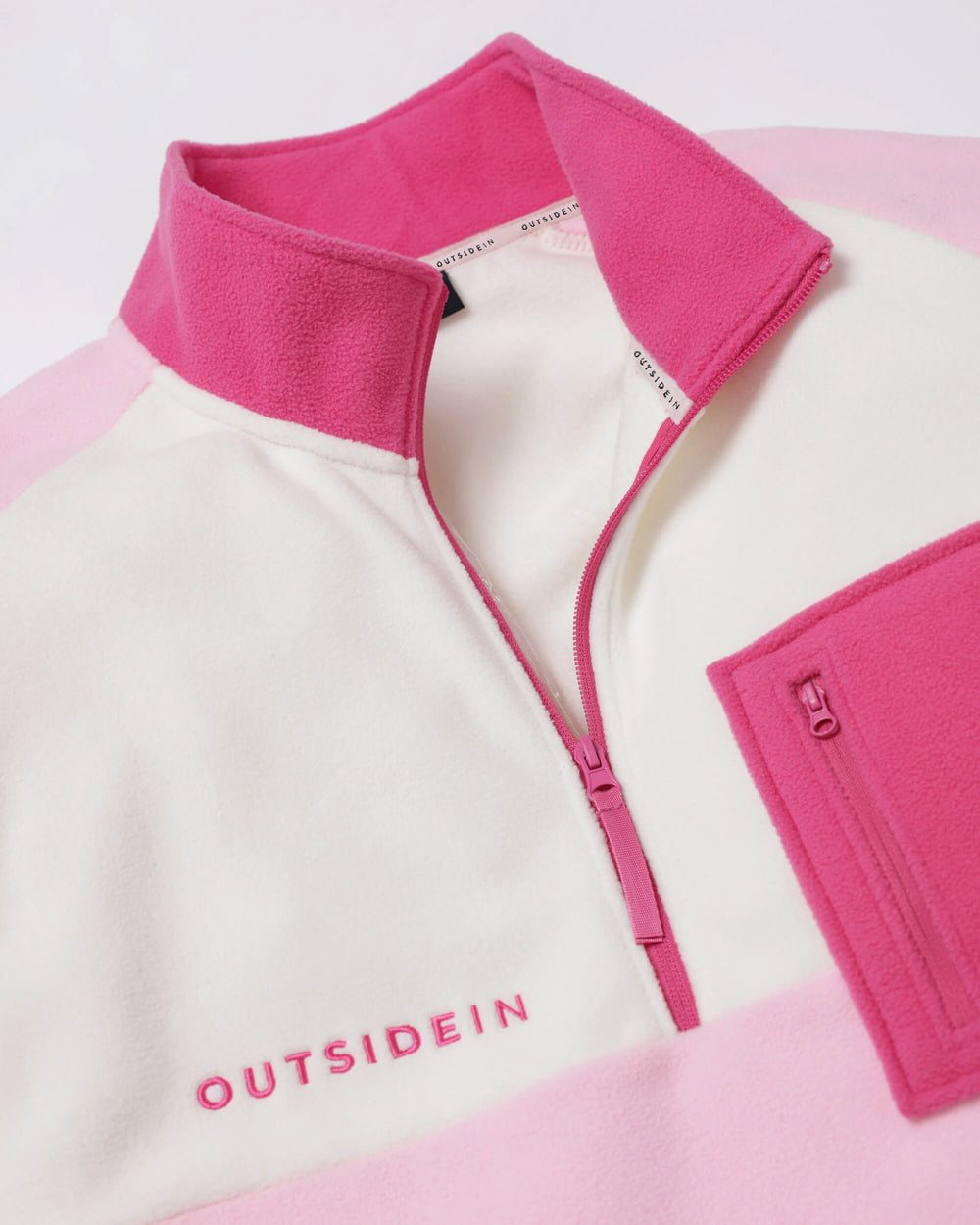 A close-up of a fleece pullover by Outsidein, featuring a white body with pink colour-blocked sections at the collar, zipper line, and lower hem. The brand name 'OUTSIDEIN' is embroidered in pink on the chest.