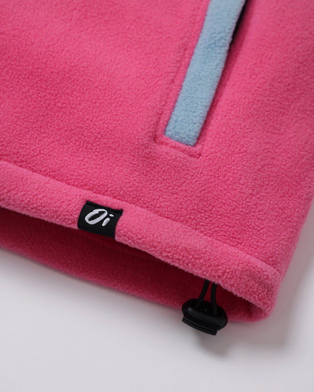 Close-up of a bright pink fleece pullover, a black fabric brand tab with white 'Oi' lettering, and a black zipper pull on a white background.