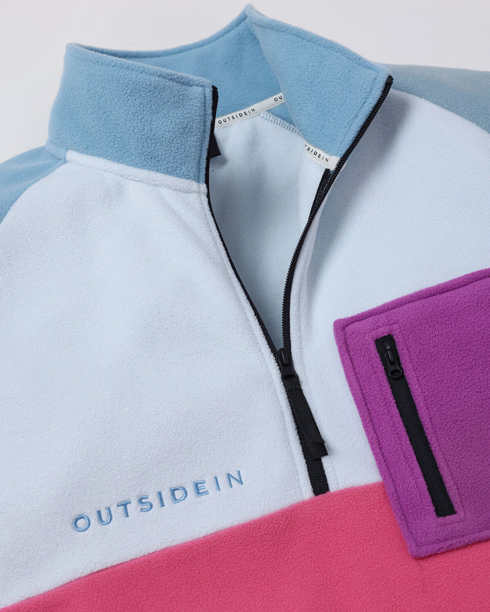 A close-up of a colour-block fleece pullover jacket by Outsidein, featuring a blue collar and chest, a bright pink bottom section and a purple zippered pocket on the right side.
