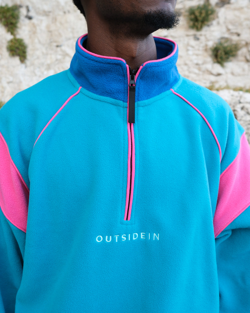 A close-up of a person wearing a bright teal quarter-zip fleece pullover with pink colour-blocked sleeves and collar trim, featuring the 'OUTSIDEIN' logo on the chest. The background shows a textured stone wall.