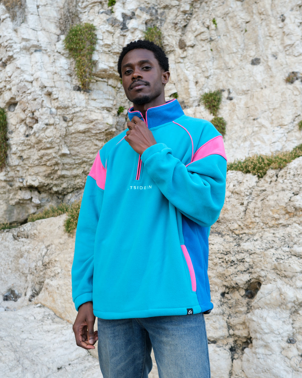 A man wearing a teal and blue quarter-zip fleece pullover with pink colour-blocked details on the shoulders and sides, standing outdoors against a light-coloured rocky cliff background.