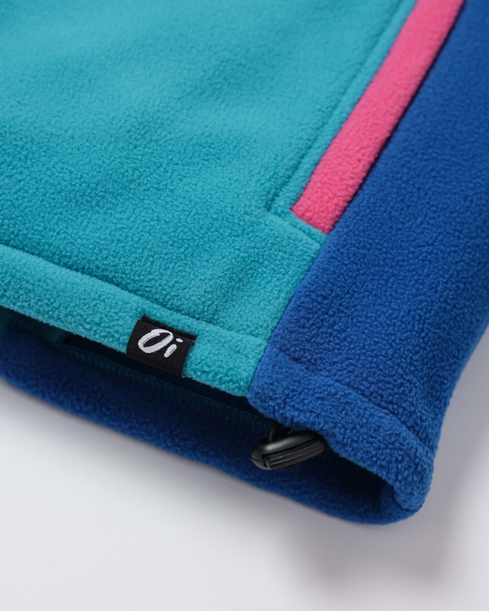 Close-up of a fleece jacket or sweatshirt corner showing a teal body, a dark blue lower section, a bright pink stripe, and a small black brand tag with white lettering.