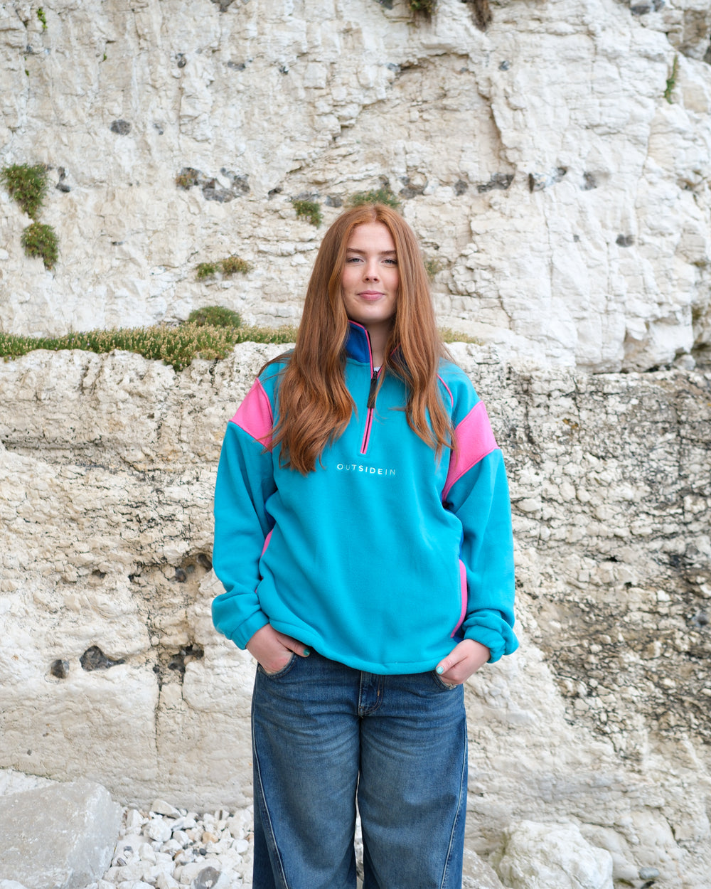 A young woman with long red hair stands in front of a white stone cliff wearing a teal and pink quarter-zip fleece pullover sweatshirt with 'OUTSIDEIN' printed in white on the chest, paired with blue jeans.