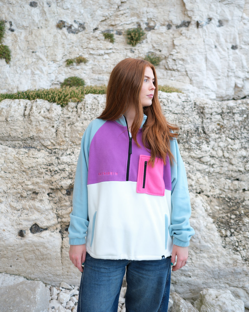 A woman with long red hair stands outdoors in front of a light-coloured stone wall, wearing a colour-blocked quarter-zip fleece pullover with light blue sleeves, a purple upper chest, a white lower body, and a bright pink pocket detail.